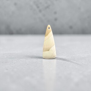 Peut inclure: Un pendentif conique de couleur claire avec un petit trou en haut. Le pendentif présente un motif en couches avec des nuances de beige et de brun clair. La surface lisse reflète la lumière, et il est posé sur une surface grise.