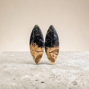 May include: Two teardrop-shaped earrings. The top portion is black with white and gold veining, and the bottom is a light brown wood grain. The earrings are set against a neutral background.