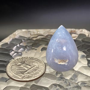 May include: A polished, pale blue gemstone in a teardrop shape. The stone has a translucent quality and a small, light-coloured inclusion. A U.S. quarter coin is next to the gemstone for size comparison. The gemstone is displayed on a textured surface.