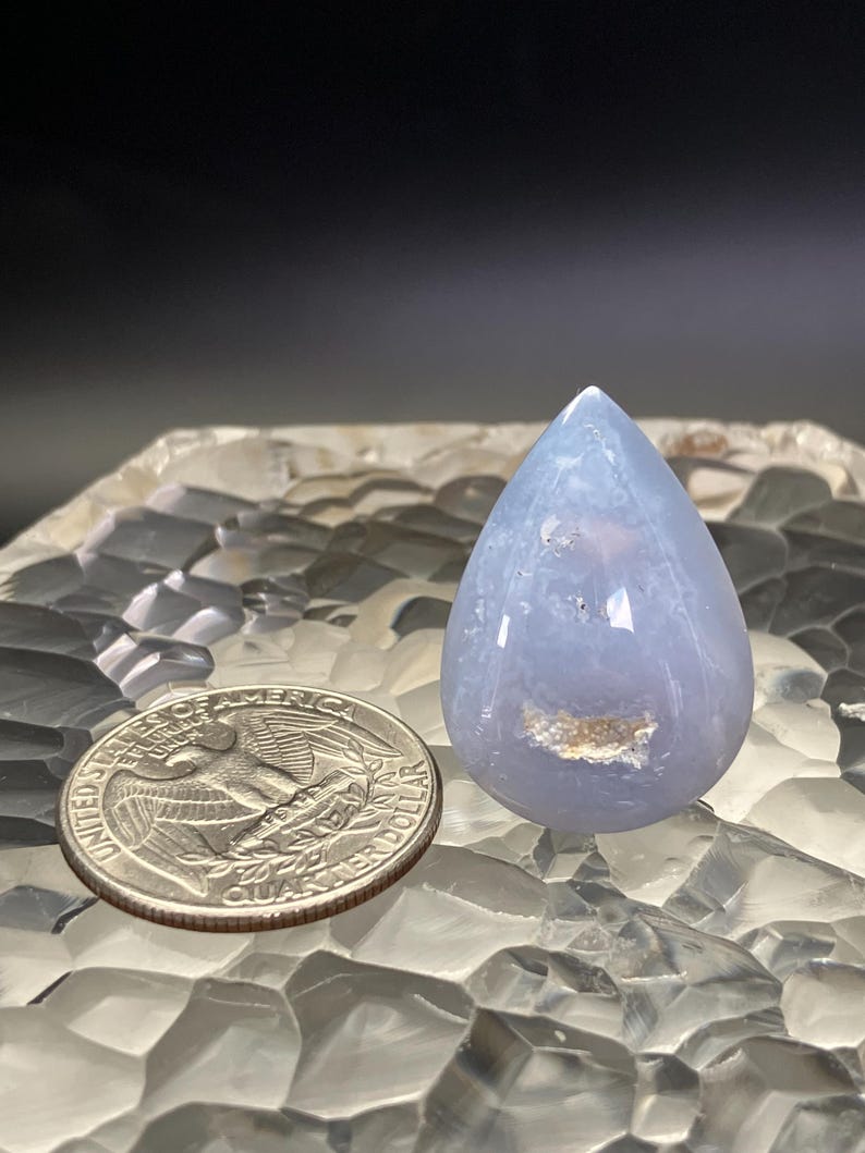 May include: A polished, pale blue gemstone in a teardrop shape. The stone has a translucent quality and a small, light-coloured inclusion. A U.S. quarter coin is next to the gemstone for size comparison. The gemstone is displayed on a textured surface.