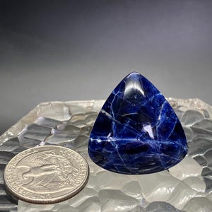 May include: A triangular blue gemstone with white veins, resembling a polished piece of lapis lazuli. The gemstone is sitting on a white, textured surface.