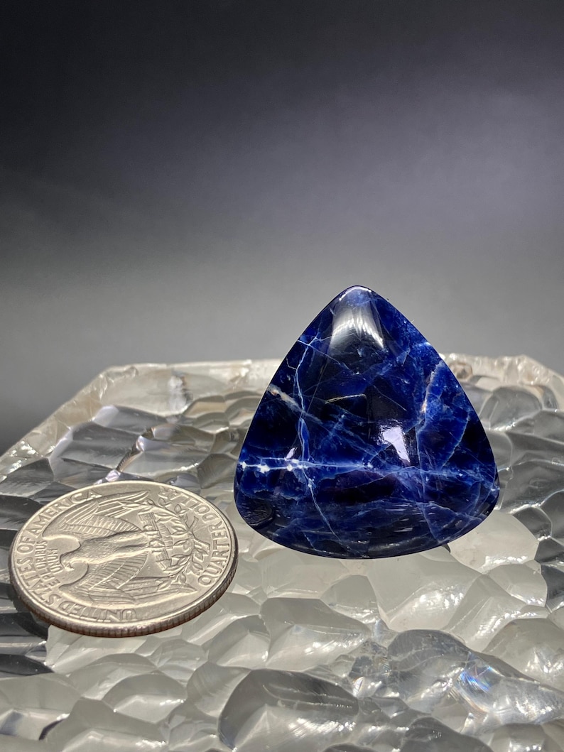 May include: A triangular blue gemstone with white veins, resembling a polished piece of lapis lazuli. The gemstone is sitting on a white, textured surface.