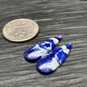 May include: Two teardrop-shaped lapis lazuli cabochons with white and blue marbling. The cabochons are approximately 1 inch long and are displayed on a dark wooden surface.