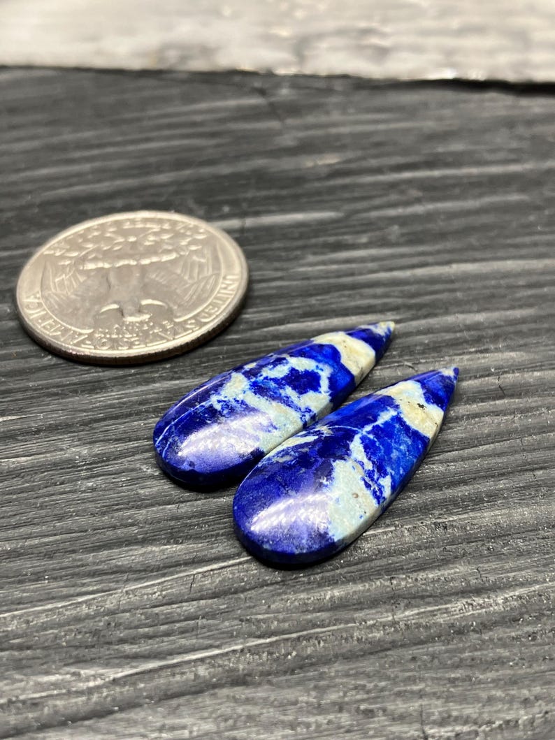 May include: Two teardrop-shaped lapis lazuli cabochons with white and blue marbling. The cabochons are approximately 1 inch long and are displayed on a dark wooden surface.