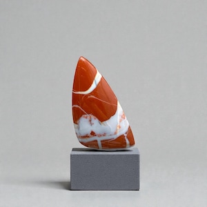 Op de afbeelding: Een gepolijste, driehoekige steen met een levendige rood-oranje basiskleur, geaderd met witte patronen. De steen is gemonteerd op een klein, grijs rechthoekig blok, wat een minimalistische presentatie creëert. De algehele esthetiek is modern en artistiek.