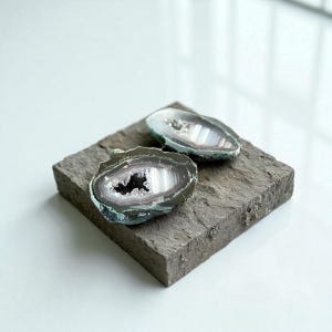 Puede incluir: Dos geodas de ágata pulidas con bandas grises, blancas y negras, expuestas sobre una base de piedra rectangular gris. Las geodas tienen un centro cristalino y un borde exterior verde oscuro. Fondo blanco.