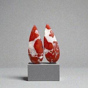 Op de afbeelding: Twee druppelvormige edelstenen met een rood-wit patroon, gepresenteerd op een grijze rechthoekige blok. De edelstenen hebben een gepolijst oppervlak en een uniek, natuurlijk ontwerp. De achtergrond is effen grijs.