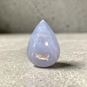 May include: A polished, teardrop-shaped gemstone in pale blue and white hues. The stone has a smooth, glossy surface with a small, light brown inclusion near the base. The gemstone is set against a grey surface.