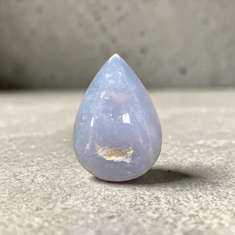May include: A polished, teardrop-shaped gemstone in pale blue and white hues. The stone has a smooth, glossy surface with a small, light brown inclusion near the base. The gemstone is set against a grey surface.