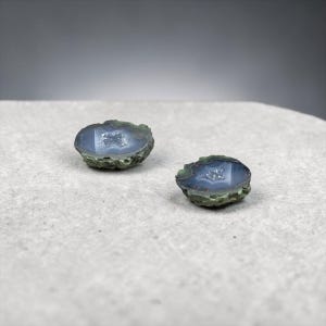 Könnte beinhalten: Zwei polierte, oval geformte Achate. Jeder Stein hat eine blau-graue Mitte mit einem kristallinen Inneren, umgeben von einer dunkelgrünen und braunen Außenschicht. Die Steine ​​werden auf einer hellgrauen Oberfläche präsentiert.