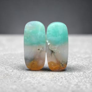 Peut inclure: Deux pierres précieuses ovales polies. Chaque pierre présente un dégradé de couleurs, avec un sommet turquoise, une transition vers le blanc et des nuances orange et marron. Les pierres ont une finition lisse et brillante sur un fond neutre.