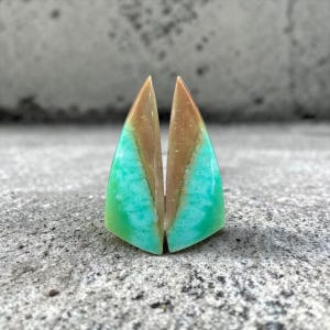 Peut inclure: Deux pierres précieuses triangulaires avec une finition polie. Les pierres présentent un dégradé de couleurs, notamment marron, turquoise et vert. Les pierres précieuses sont exposées sur une surface grise texturée.