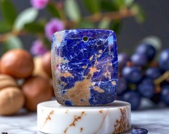 May include: A rectangular, blue and brown gemstone pendant with a drilled hole. The stone is displayed on a white marble stand. The background includes blurred flowers and grapes.