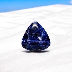 May include: A triangular blue gemstone with white veins, sitting on a white surface.