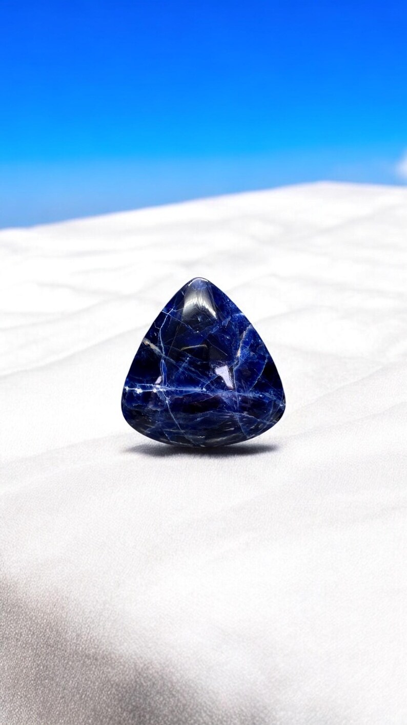 May include: A triangular blue gemstone with white veins, sitting on a white surface.