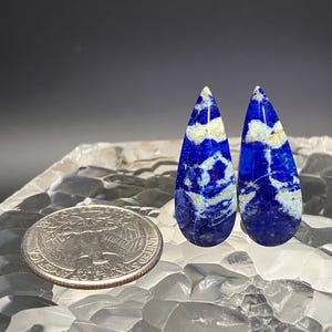 May include: Two teardrop-shaped lapis lazuli cabochons with a white and blue marbled pattern. The cabochons are about the size of a US quarter.