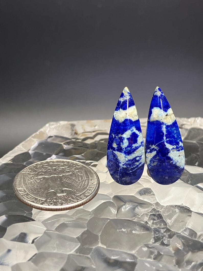 May include: Two teardrop-shaped lapis lazuli cabochons with a white and blue marbled pattern. The cabochons are about the size of a US quarter.