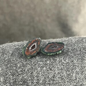 May include: Two polished agate slices. Each slice has a dark red center with a crystalline interior, surrounded by a band of orange and a green outer edge. The stones are displayed on a gray, textured surface.