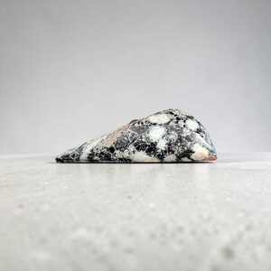 Peut inclure: Une pierre polie de forme triangulaire présentant un motif complexe d'inclusions minérales noires, blanches et rose pâle. La surface offre une texture lisse et brillante avec un aspect tacheté, révélant des amas cristallins distincts.