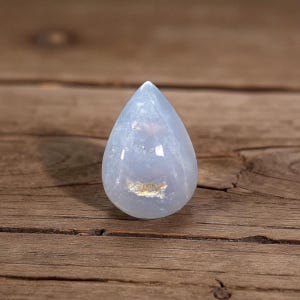 May include: A pale blue, teardrop-shaped gemstone with a polished surface. The stone has a translucent quality, with subtle variations in colour and texture. The gemstone is displayed on a wooden surface.