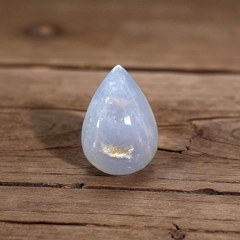 May include: A pale blue, teardrop-shaped gemstone with a polished surface. The stone has a translucent quality, with subtle variations in colour and texture. The gemstone is displayed on a wooden surface.