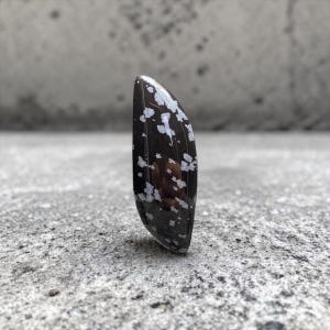 Peut inclure: Une pierre d'obsidienne flocon de neige polie en forme de goutte d'eau. La pierre noire présente des inclusions blanches en forme de flocons de neige. La pierre est présentée sur une surface grise et texturée.
