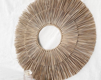 May include: A large, round, natural woven wall hanging with a circular opening in the center. The wall hanging is made of light brown, straw-like material and has a sunburst design.