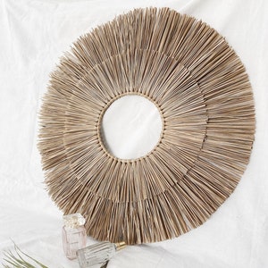 Puede incluir: Un gran tapiz de pared redondo tejido de forma natural con una abertura circular en el centro. El tapiz de pared está hecho de un material de paja marrón claro y tiene un diseño de rayos de sol.