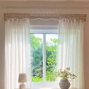 K&ouml;nnte beinhalten: Eine Kommode aus hellem Holz mit sechs Schubladen und abgerundeten Ecken, darauf eine kleine plissierte Lampe und eine gerippte Vase mit wei&szlig;en Blumen. Dar&uuml;ber befindet sich ein Fenster mit halbtransparenten wei&szlig;en Vorh&auml;ngen und einem gewebten, gebogten Volant mit der Aufschrift MADEBYIND.