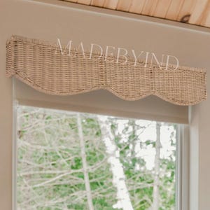 Op de afbeelding: Een slaapkamer met een raam met een geweven valance en uitzicht op bomen. Een stapel gevouwen beige handdoeken ligt op een bed met een gestreepte deken. De tekst "MADEBYIND" is zichtbaar.