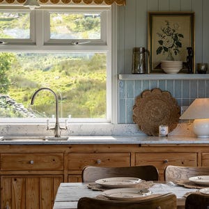 Puede incluir: Una cocina con armarios de madera natural, una encimera blanca moteada y un grifo de acero inoxidable. Una mesa de comedor con sillas de madera y platos de cer&aacute;mica se encuentra en primer plano. Una ventana da a un paisaje verde.