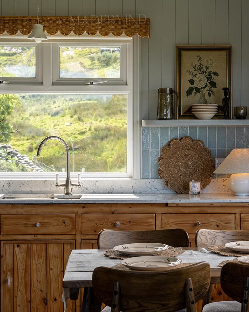 Puede incluir: Una cocina con armarios de madera natural, una encimera blanca moteada y un grifo de acero inoxidable. Una mesa de comedor con sillas de madera y platos de cer&aacute;mica se encuentra en primer plano. Una ventana da a un paisaje verde.
