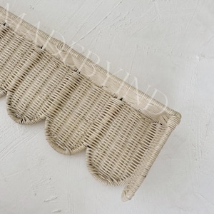 Puede incluir: Un estante de pared de rat&aacute;n tejido en color beige claro con un borde superior recto y un borde inferior festoneado decorativo. El estante tiene una textura natural y un patr&oacute;n semicircular en la parte inferior, hecho de material de junco fino.