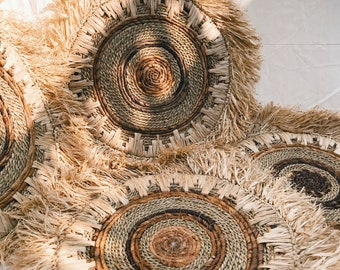 Puede incluir: Tres piezas redondas de decoración de pared tejidas con un borde de flecos. Las piezas tejidas tienen una gama de colores naturales marrones y beige.