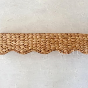 May include: A woven, natural-toned, scalloped-edge shelf made of rattan or seagrass. The shelf is a light brown color and has a wavy edge.