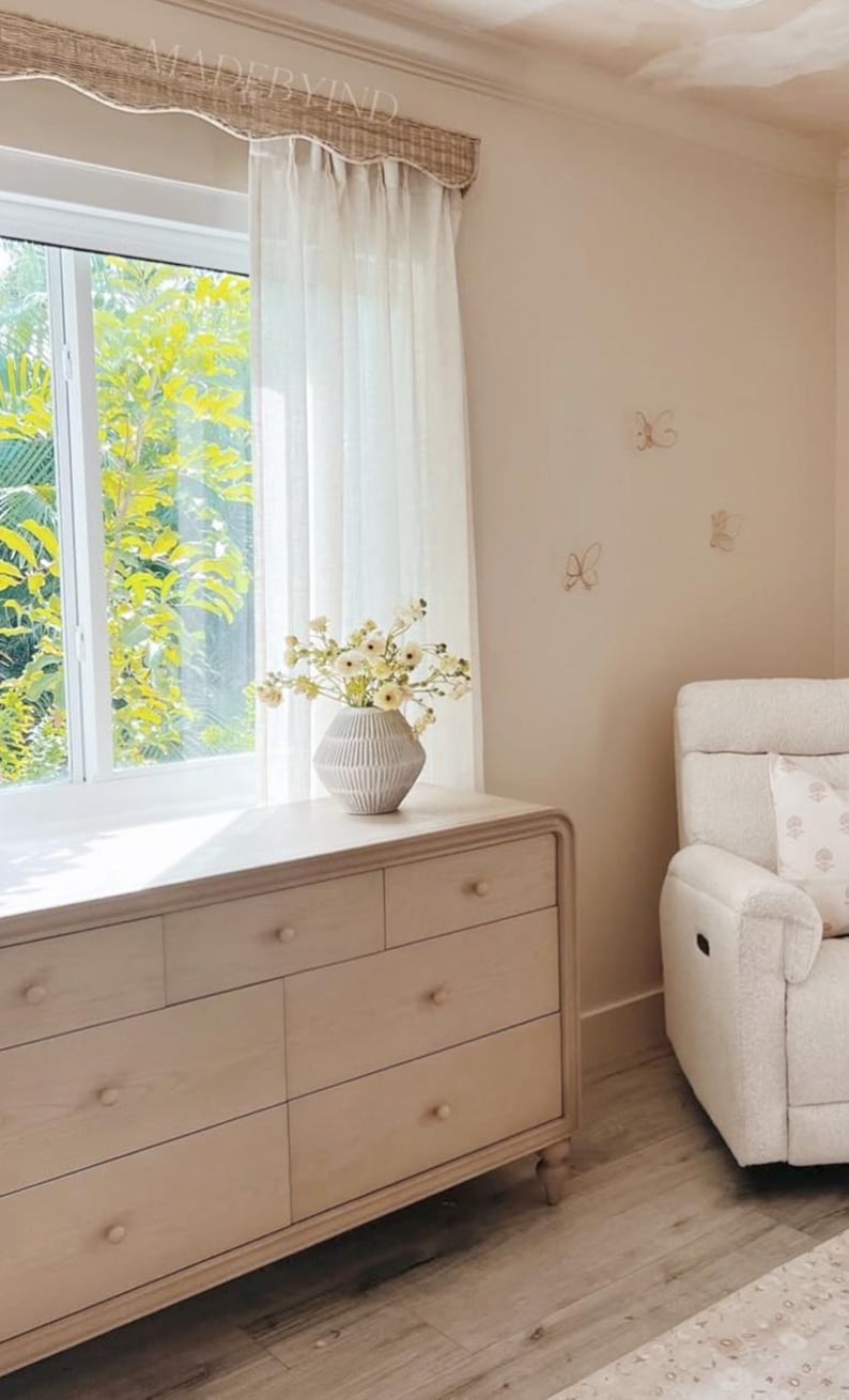 K&ouml;nnte beinhalten: Eine helle Holzkommode mit sechs Schubladen und runden Kn&ouml;pfen steht unter einem Fenster mit wei&szlig;en Vorh&auml;ngen. Eine gerippte wei&szlig;e Vase mit wei&szlig;en Blumen steht darauf. Drei kleine Schmetterlings-Wanddekorationen sind an der beigen Wand.