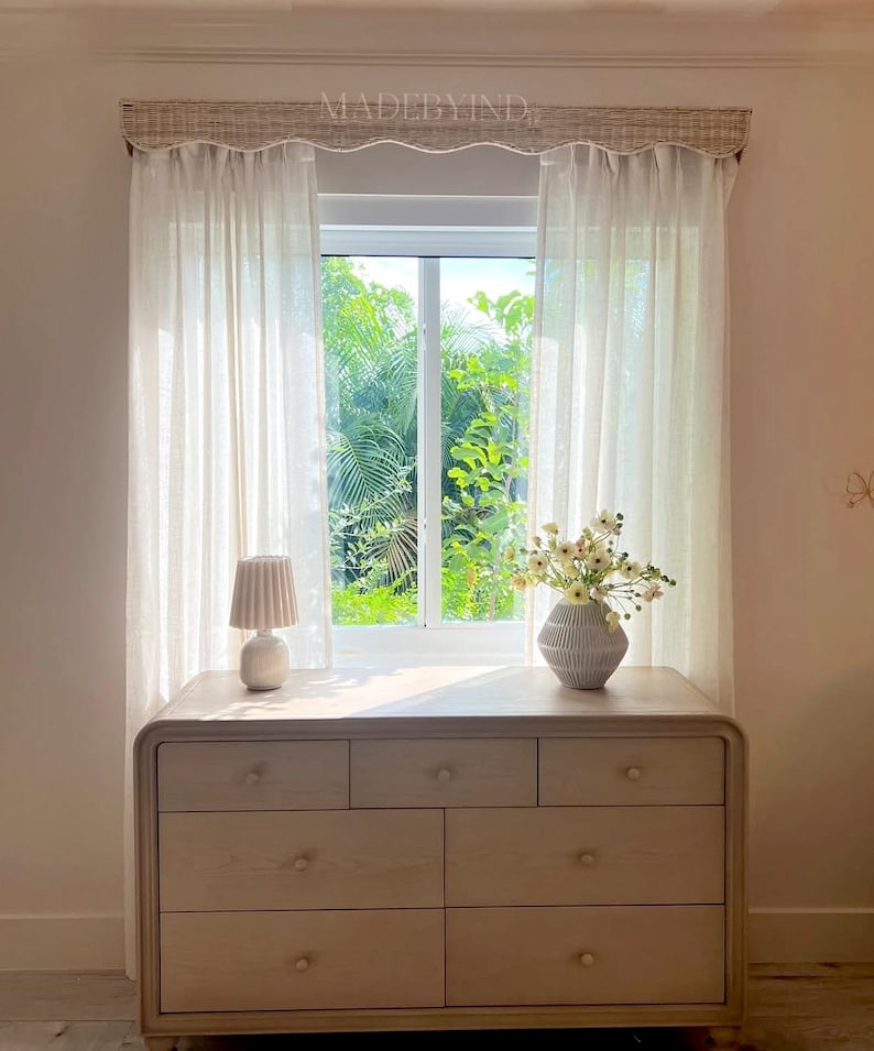K&ouml;nnte beinhalten: Eine Kommode aus hellem Holz mit sechs Schubladen und abgerundeten Ecken, darauf eine kleine plissierte Lampe und eine gerippte Vase mit wei&szlig;en Blumen. Dar&uuml;ber befindet sich ein Fenster mit halbtransparenten wei&szlig;en Vorh&auml;ngen und einem gewebten, gebogten Volant mit der Aufschrift MADEBYIND.