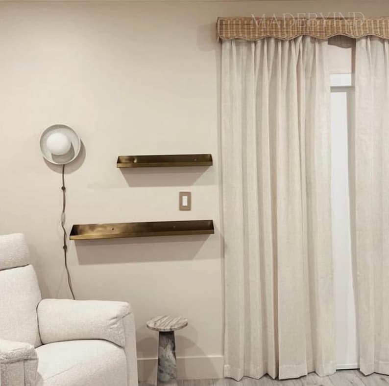 K&ouml;nnte beinhalten: Zwei horizontale Wandregale aus Metall mit dunkelbronzefarbenem Finish an einer hellbeigen Wand. Eine kreisf&ouml;rmige Wandleuchte mit wei&szlig;er Kugellampe, ein hell gepolsterter Sessel, ein kleiner Beistelltisch aus Marmor und bodenlange Vorh&auml;nge.