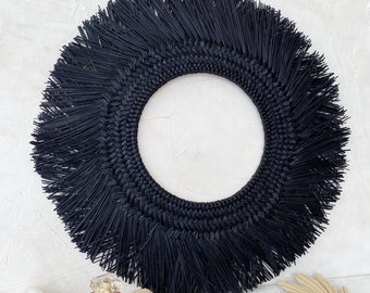 Puede incluir: Colgante de pared tejido negro con borde de flecos. El colgante es redondo y tiene un aspecto natural y rústico.