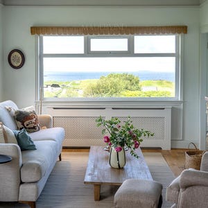 Puede incluir: Una sala de estar con un sof&aacute; gris claro con cojines, una mesa de centro de madera con un jarr&oacute;n de flores rosas, un taburete a juego y una ventana grande con una cenefa festoneada que da a un paisaje costero con vistas al mar.