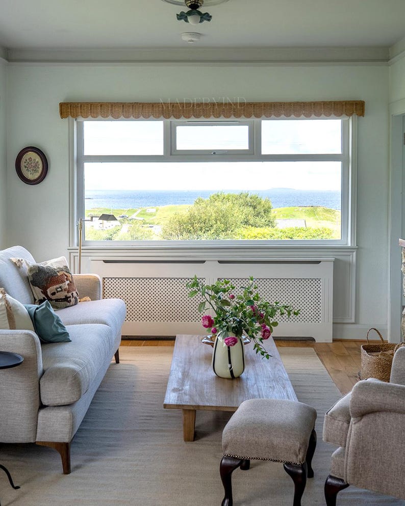 Puede incluir: Una sala de estar con un sof&aacute; gris claro con cojines, una mesa de centro de madera con un jarr&oacute;n de flores rosas, un taburete a juego y una ventana grande con una cenefa festoneada que da a un paisaje costero con vistas al mar.