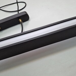 May include: A modern, linear pendant light fixture. The fixture features a dark wood frame with a recessed, bright white light source. Black cords and a mounting plate are also visible.