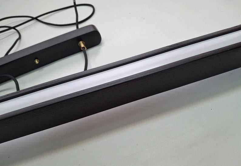 May include: A modern, linear pendant light fixture. The fixture features a dark wood frame with a recessed, bright white light source. Black cords and a mounting plate are also visible.
