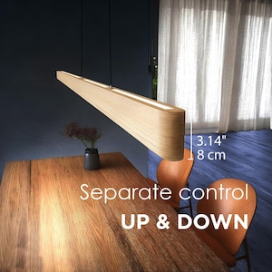 May include: A modern, light wood-toned pendant light fixture, 3.14 inches (8 cm) in height, hangs above a wooden table. The fixture has a built-in light and is suspended by thin black cords. The text "Separate control UP & DOWN" is visible.