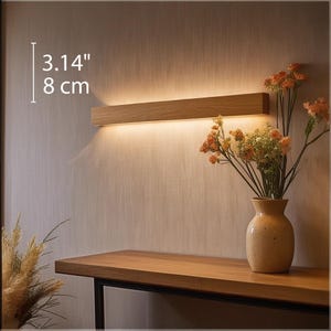 Könnte beinhalten: Eine Wandleuchte aus Holz mit warmweißem Licht. Die Leuchte ist 3,14 Zoll lang und 8 Zentimeter breit. Die Leuchte ist an einer Wand montiert, vor der sich ein Holztisch befindet. Auf dem Tisch steht eine Vase mit Trockenblumen.