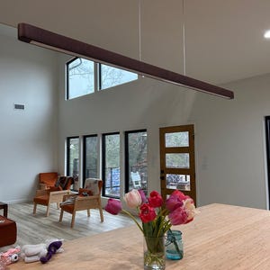 May include: A modern, rectangular, brown wooden pendant light fixture suspended from the ceiling. The light is above a wooden countertop with a vase of tulips and a blue jar. The room has large windows and two orange armchairs.