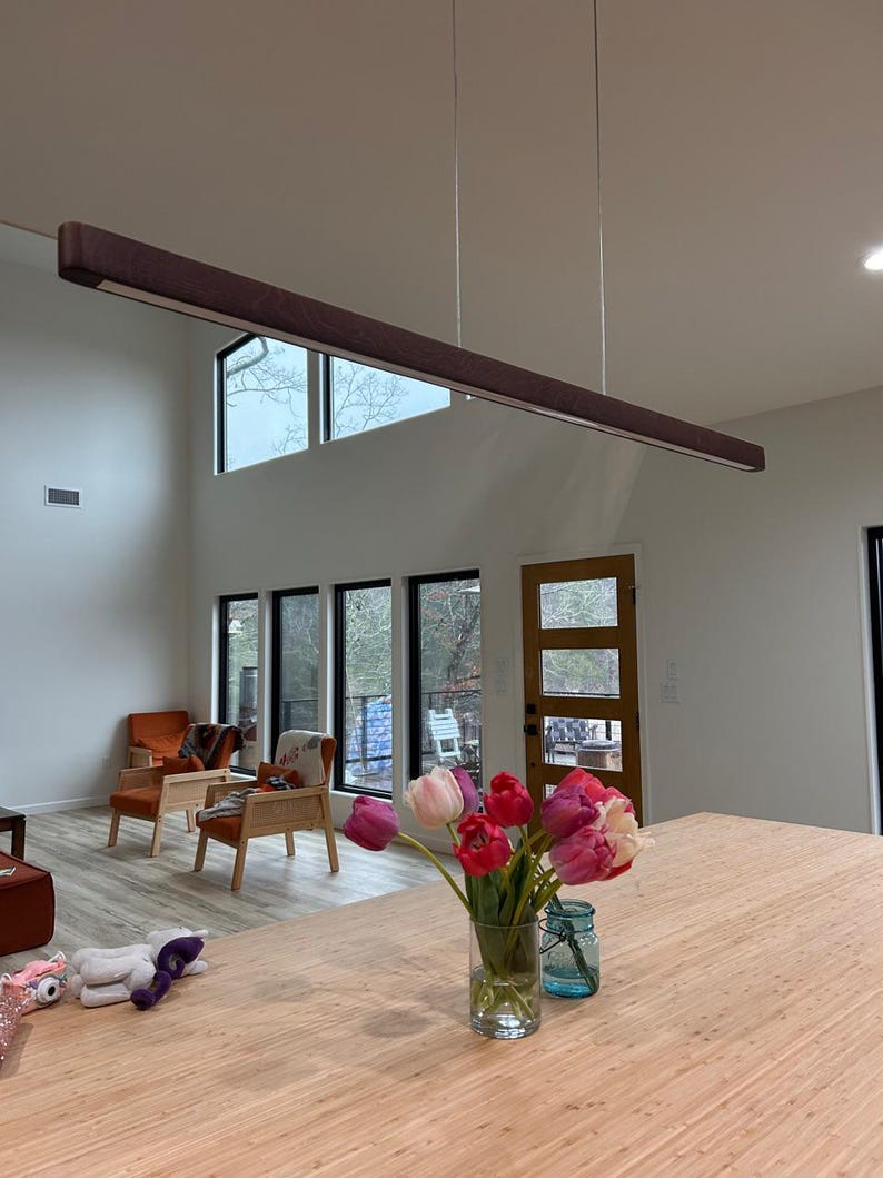 May include: A modern, rectangular, brown wooden pendant light fixture suspended from the ceiling. The light is above a wooden countertop with a vase of tulips and a blue jar. The room has large windows and two orange armchairs.