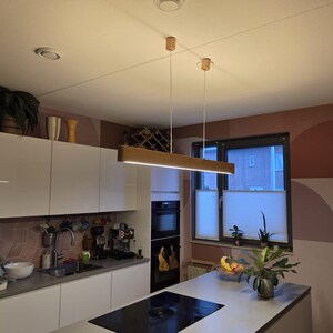 May include: A modern kitchen with white cabinets, a gray countertop, and a black stovetop. A rectangular wooden light fixture hangs from the ceiling. The room has a window with blinds and a decorative wall with geometric shapes.