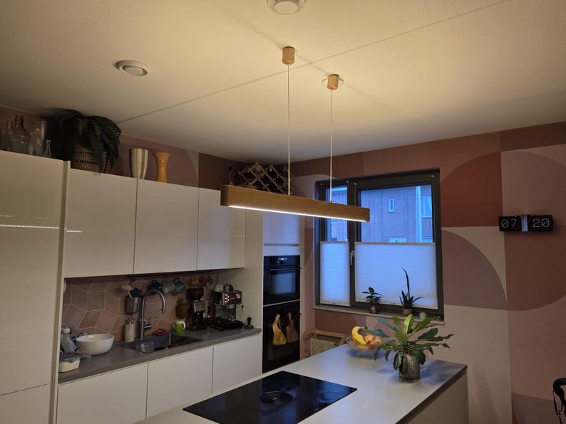 May include: A modern kitchen with white cabinets, a gray countertop, and a black stovetop. A rectangular wooden light fixture hangs from the ceiling. The room has a window with blinds and a decorative wall with geometric shapes.