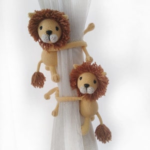 May include: Two crocheted lion curtain tiebacks in beige and brown with white faces. The tiebacks are holding up a sheer white curtain.
